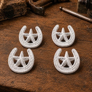 May include: A set of four white, distressed-finish metal decorative accents, each shaped like a horseshoe with a five-pointed star centered within the arch. The pieces feature a textured, weathered appearance with visible dark undertones.