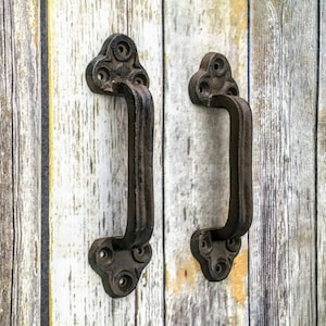 May include: Two black cast iron door handles with a decorative design on a weathered wood background.
