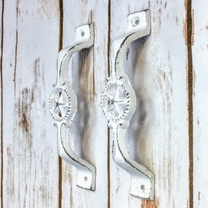 May include: Two white cast iron door pulls with a star design. The pulls are mounted on a white wood background.