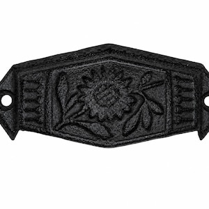 May include: A black cast iron drawer pull with a textured finish. The hardware features a central embossed sunflower design with leaves, flanked by vertical line patterns on each side. It has a diamond-shaped frame with two screw holes.