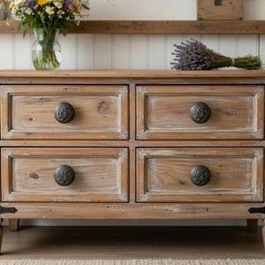 Puede incluir: Cómoda de madera marrón claro con cuatro cajones, cada uno con un pomo oscuro y ornamentado. La cómoda tiene un acabado envejecido y detalles de esquina de metal negro. Un ramo de flores y lavanda encima.