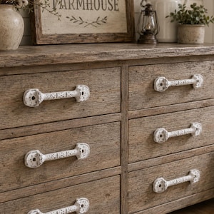 May include: A wooden dresser featuring six drawers with a weathered brown finish and textured grain. Each drawer is equipped with a horizontal, distressed white metal handle that has a decorative, ornate pattern and circular end plates.