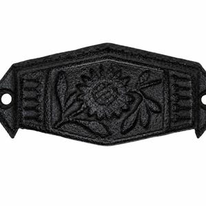 Puede incluir: Un tirador de cajón de hierro fundido negro con acabado texturizado. El herraje presenta un diseño central en relieve de un girasol con hojas, flanqueado por patrones de líneas verticales a cada lado. Marco en forma de diamante con dos orificios.