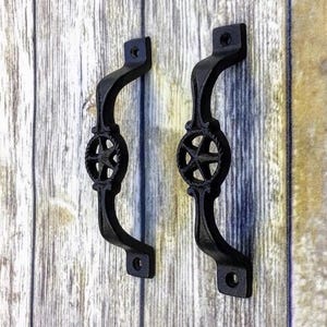 May include: A pair of black cast iron door handles. Each handle features a curved design with a star motif at the centre, encircled by a rope-like border. They are mounted on a wooden surface.