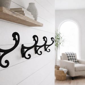 May include: A row of four black metal coat hooks mounted on a white shiplap wall. The hooks have a decorative, scrolled design. A wooden shelf with decor is above the hooks. The room has a neutral color palette.