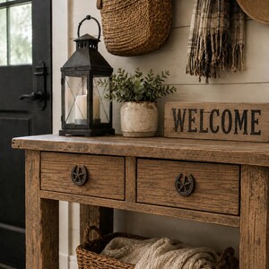 May include: A wooden console table with two drawers featuring horseshoe-shaped pulls. On top sits a black metal lantern with a candle, a small potted plant, and a wooden sign with the word WELCOME. A woven basket with a textile sits on the lower shelf.