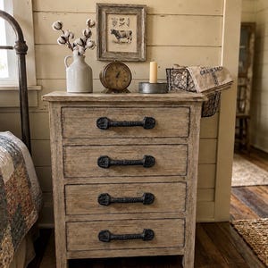 May include: A light-colored wooden four-drawer chest featuring dark, textured metal bar handles on each drawer. The chest has a weathered finish and is positioned next to a bed with a patchwork quilt, topped with a white pitcher, a clock, and a framed print.