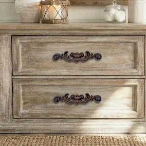 May include: A distressed, light-brown wooden nightstand with two drawers. The drawers have ornate, dark-brown metal handles. Decorative items, including a candle in a glass holder, sit on top of the nightstand.