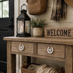 Peut inclure: Une console en bois avec deux tiroirs dotés de poignées en forme de fer à cheval et d'étoile blanches. La table porte une lanterne en métal noir avec une bougie, une plante verte en pot et un panneau en bois avec le mot WELCOME en noir.