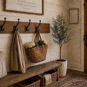 May include: A wall-mounted wooden coat rack featuring four black metal hooks. The rack is mounted on a horizontal wooden board. A tan jacket hangs on one hook, and a woven basket containing white flowers hangs on another hook.