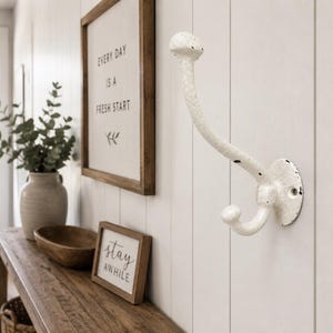 May include: A white, double-pronged wall hook with a textured, distressed finish. The metal hook features a curved upper arm and a smaller lower hook, mounted to a circular base with two visible screw holes for attachment to a vertical surface.