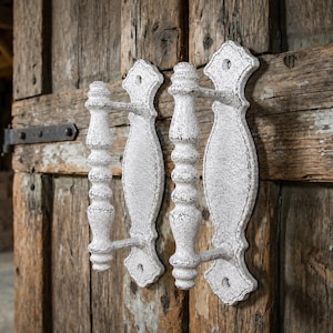 May include: A pair of white door handles with a distressed, crackled finish. Each handle features a central vertical grip with decorative turned detailing and is mounted on an elongated, shaped backplate with screw holes at the top and bottom.
