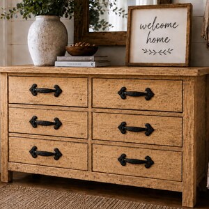 Puede incluir: Una cómoda de madera de color marrón claro con seis cajones, cada uno con un tirador de metal negro. La cómoda tiene un acabado texturizado y desgastado, y está decorada con un jarrón blanco, un cuenco pequeño, dos libros y un letrero con el texto welcome home.