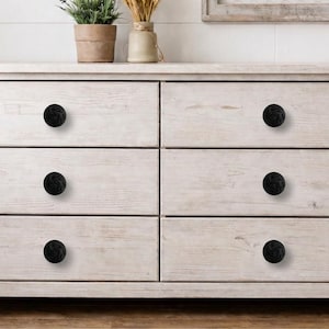 May include: A white wooden dresser with six drawers, each fitted with a round, black knob. The dresser has a distressed finish, and the knobs provide a contrasting accent. The overall style is rustic and farmhouse-inspired.
