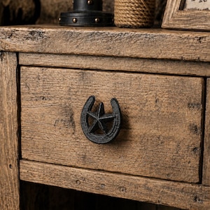 Puede incluir: Un cajón de madera rústica con un prominente tirador de hierro fundido negro en forma de herradura con una estrella de cinco puntas en el centro. La madera tiene un acabado marrón texturizado y desgastado con vetas visibles.