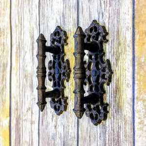 May include: Two black metal drawer pulls with ornate designs. The pulls have a decorative, scroll-like design and a central handle.
