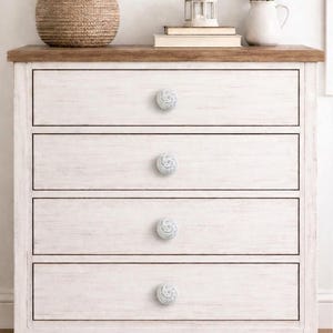 May include: A white painted wooden dresser with four drawers, each with a decorative, swirl-patterned knob. The top is a contrasting brown wood. A woven basket of white flowers and other decorative items sit on top.