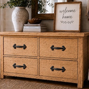 Puede incluir: Una cómoda de madera de color marrón claro con cuatro cajones y tiradores de metal negro. El mueble tiene un acabado texturizado y se apoya sobre una base plana. Un letrero cuadrado de madera con el texto "welcome home" y un diseño de hojas descansa encima.