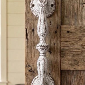 May include: A vertical door handle with a distressed white finish and dark brown undertones. The handle features a central spindle design with two circular mounting plates at the top and bottom, each decorated with intricate radial patterns and screw holes.