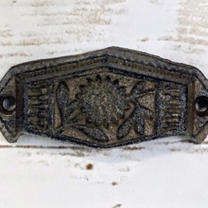 May include: A cast iron drawer pull with a floral design. The pull is a dark gray color and has a slightly aged look.