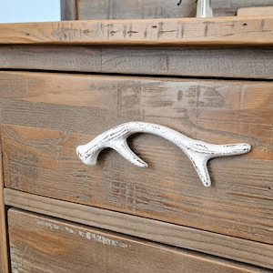 May include: A wooden dresser drawer featuring a white, distressed-finish antler-shaped handle. The drawer front is constructed from light brown, weathered wood with a visible grain pattern and horizontal planks, showcasing a rustic aesthetic.