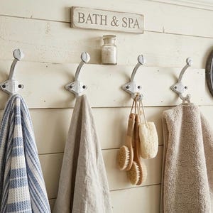 Op de afbeelding: Rustieke badkamerwanddecoratie met een houten bord met de tekst "BATH & SPA". Daaronder hangen vier witte metalen haken met handdoeken en badaccessoires. Rechts is een kleine ronde spiegel.