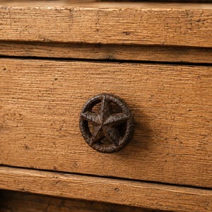 May include: A wooden drawer front featuring a prominent, circular, dark brown, textured metal knob with a five-pointed star design centered in the middle. The wood displays a natural, light brown grain with visible knots and a rough, weathered texture.