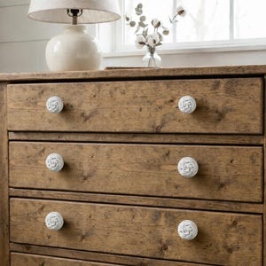 Puede incluir: Cómoda rústica de madera con seis cajones, cada uno adornado con un pomo blanco estampado. La cómoda es de madera marrón claro y tiene una lámpara encima. La habitación tiene una paleta de colores neutros.