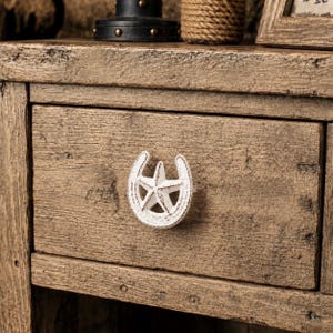 May include: A white, distressed-finish metal drawer knob shaped like a horseshoe with a five-pointed star in the center. The knob is mounted on a single wooden drawer front featuring a rough, natural wood grain texture and brown tones.