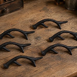 May include: A set of six black cast iron drawer pulls shaped like deer antlers. Each handle features a curved, textured design with a single branch point, providing a sturdy grip for cabinets, cupboards, or furniture doors in a home setting.