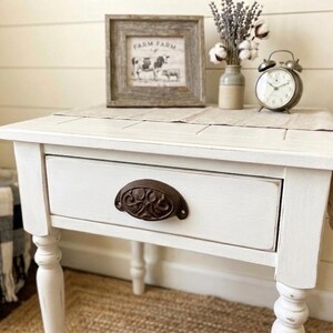Peut inclure: Une table de chevet blanche vieillie avec un tiroir et une poign&eacute;e en m&eacute;tal fonc&eacute;. La table a des pieds tourn&eacute;s et une petite image encadr&eacute;e sur le dessus, avec un vase de fleurs s&eacute;ch&eacute;es et un r&eacute;veil vintage. La table est pos&eacute;e sur un tapis tiss&eacute;.