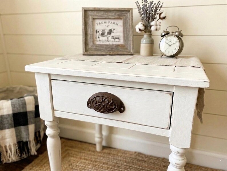 Peut inclure: Une table de chevet blanche vieillie avec un tiroir et une poign&eacute;e en m&eacute;tal fonc&eacute;. La table a des pieds tourn&eacute;s et une petite image encadr&eacute;e sur le dessus, avec un vase de fleurs s&eacute;ch&eacute;es et un r&eacute;veil vintage. La table est pos&eacute;e sur un tapis tiss&eacute;.