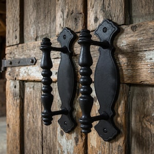 Puede incluir: Dos manijas de puerta de metal negro con un agarre en forma de huso y una placa decorativa con bordes festoneados. Las manijas están montadas verticalmente en una puerta de madera de color marrón claro desgastada con veta visible.