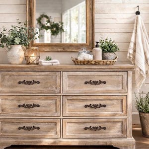 Könnte beinhalten: Eine hellfarbene Holzkommode mit sechs Schubladen, verzierten dunklen Metallgriffen und einem Used-Look. Die Kommode ist mit dekorativen Gegenständen wie Pflanzen, einem Spiegel und einem geflochtenen Tablett versehen. Ein geflochtener Korb und ein Metalleimer mit einer Pflanze sind ebenfalls zu sehen.