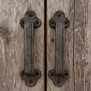 May include: Two dark, textured, cast iron door handles mounted vertically on weathered, grey wooden planks. Each handle features a decorative, clover-shaped base plate secured with four black screws, showcasing a rugged, aged metal finish.