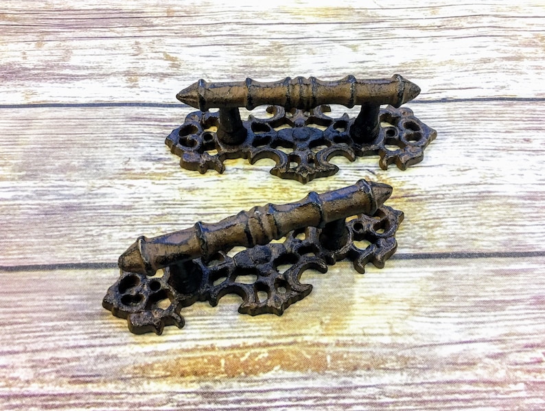 Puede incluir: Dos tiradores de caj&oacute;n de hierro fundido ornamentados con un dise&ntilde;o de desplazamiento decorativo y un acabado marr&oacute;n. Los tiradores tienen una base rectangular y una manija corta y recta.