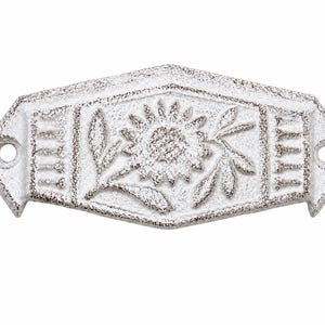 Puede incluir: Un tirador de cajón de metal fundido blanco con un acabado texturizado y desgastado. La pieza rectangular presenta un diseño central en relieve de girasol con hojas y patrones de líneas verticales en los lados, incluyendo dos orificios.