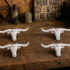 Peut inclure: Un ensemble de quatre patères murales blanches en forme de têtes de bœuf longhorn. Chaque crochet présente une finition vieillie et usée avec une texture sombre visible, de longues cornes incurvées et un mécanisme de fixation au dos.