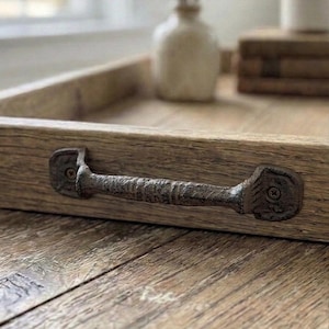 Peut inclure: Un plateau de service en bois rustique avec une poignée en métal foncé. Le plateau est en bois clair avec un grain visible et repose sur une surface en bois. Un petit vase et des objets décoratifs sont en arrière-plan.