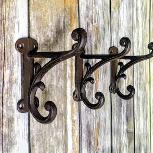 May include: Ornate, cast iron wall hooks in a dark brown finish. The hooks feature a decorative scroll design and are mounted on a wooden background. Ideal for hanging coats, hats, or other items.