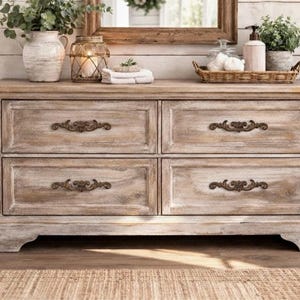 May include: A distressed wooden dresser with four drawers, each adorned with ornate bronze-colored handles. The dresser is light brown with white accents, and is topped with decorative items, including a vase, candle holder, and a small tray.