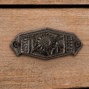 May include: A dark grey, cast metal drawer pull with a hexagonal shape and a central embossed floral design. The hardware features two small screws on the left and right sides, securing the piece to a light brown, horizontal wood grain surface.