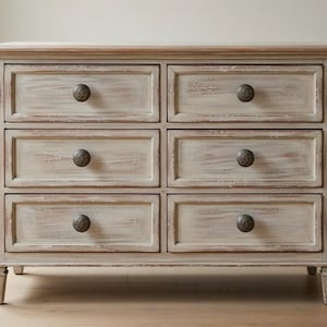 Puede incluir: Cómoda de seis cajones con acabado blanquecino envejecido. Los cajones tienen pomos redondos y oscuros y están enmarcados con un borde de madera más oscuro. La cómoda se apoya sobre patas cortas y decorativas y tiene una tapa de color claro.