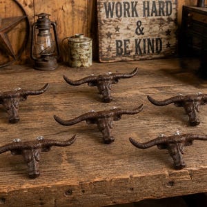 Puede incluir: Un conjunto de cinco ganchos de pared de hierro fundido en color marrón oscuro, cada uno con forma de cabeza de toro longhorn con cuernos curvos prominentes. Estos ganchos de metal tienen un acabado texturizado y orificios de montaje.