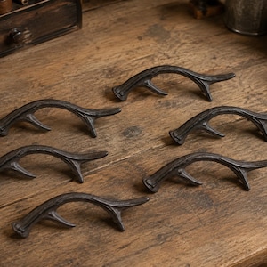 Peut inclure: Un ensemble de six poignées de placard marron foncé en forme de bois de cerf. Chaque poignée présente un design incurvé et texturé avec une seule pointe s'étendant sur le côté, imitant l'apparence d'un bois naturel en métal massif.