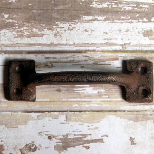 May include: A rustic black cast iron drawer pull with a rectangular shape and a slightly curved handle. The pull has two holes for mounting.