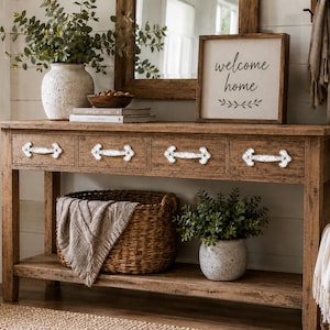 Puede incluir: Una consola de madera rústica con cuatro cajones y tiradores de metal blanco ornamentados. El estante inferior contiene una cesta tejida con una manta beige y una planta verde. Encima hay un letrero con el texto welcome home.