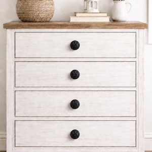May include: A white wooden dresser with four drawers, each with a black, textured knob. The top is a natural wood color. A woven basket of white flowers, a lantern, and a vase of lavender sit on top.