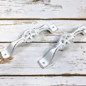 May include: Two white cast iron drawer pulls with a star design. The pulls are on a white wood background.