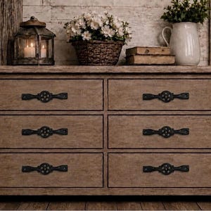 May include: A rustic wooden dresser with six drawers, each featuring ornate black hardware. The dresser is topped with decorative items, including a lantern, flowers, and a pitcher, creating a warm, inviting aesthetic.